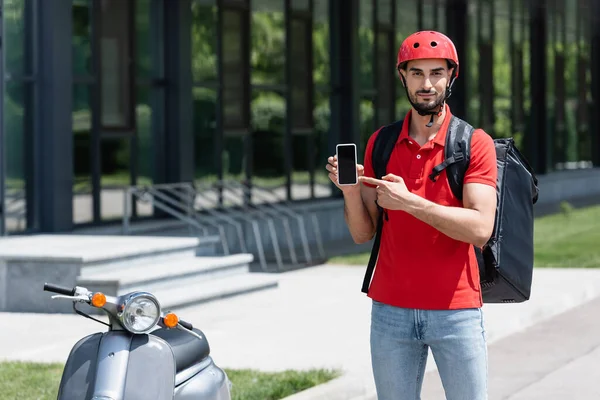 戴头盔和热身背包的阿拉伯信使指着户外滑板车旁边装有空白屏幕的智能手机 — 图库照片