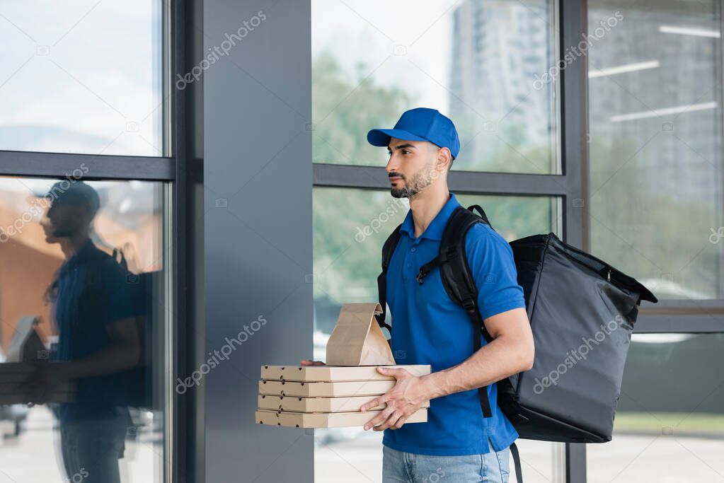 Arabian deliveryman holding paper bag and pizza boxes near building 