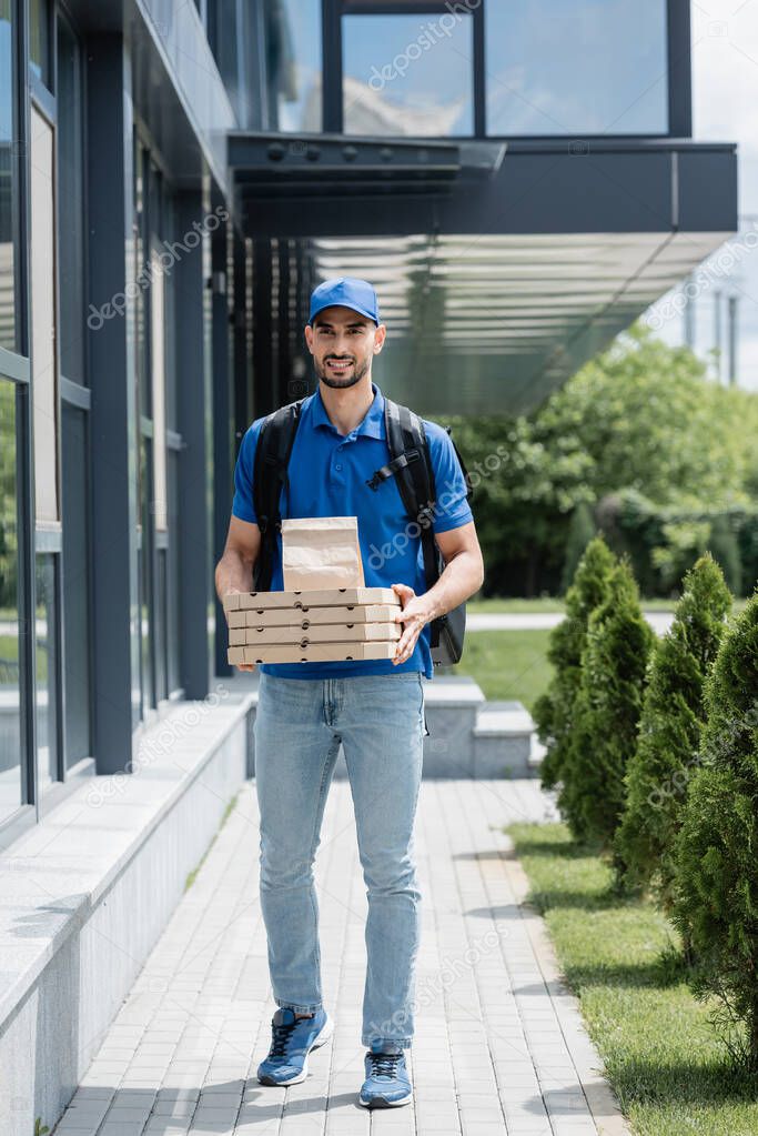 Positive muslim courier holding paper bag and pizza boxes near building 