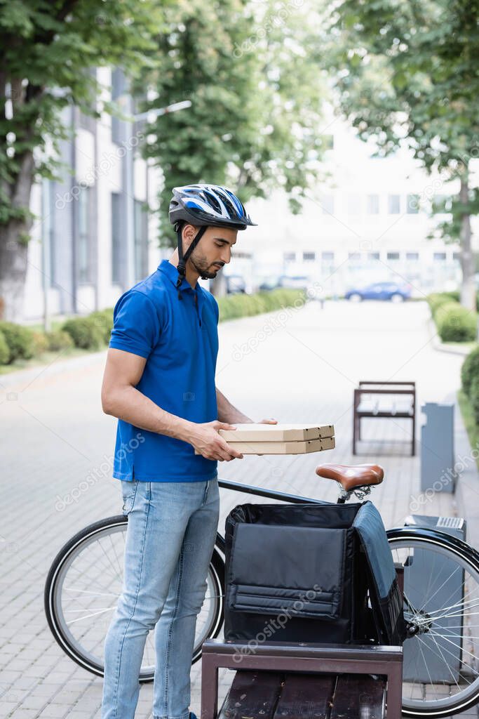 Side view of arabian deliveryman holding pizza boxes near thermo backpack and bike outdoors 