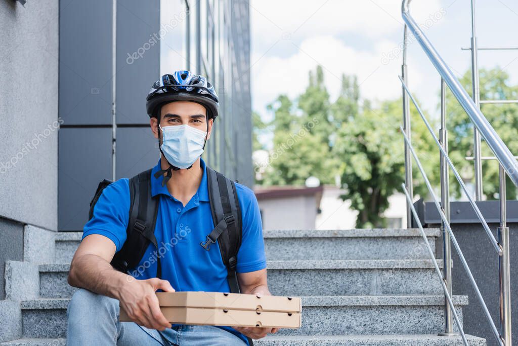 Muslim deliveryman in helmet and medical mask holding pizza boxes on stairs of building 