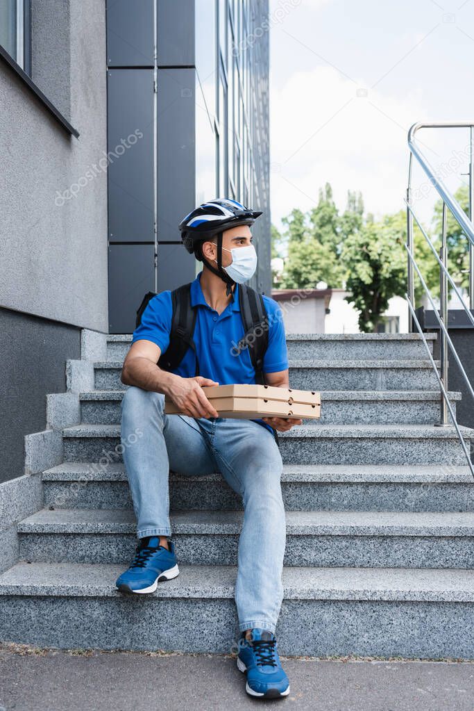 Arabian courier in medical mask and safety helmet holding pizza boxes on stairs outdoors 