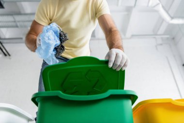 Low angle view of man in latex gloves holding plastic bags near trash can with recycle sign  clipart