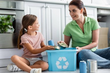 Smiling woman and daughter holding tin cans near box with recycle sign in kitchen  clipart