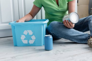 Cropped view of woman holding tin can near box with recycle sign on floor  clipart