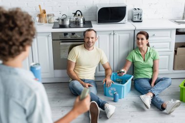 Gülümseyen aileler geri dönüşüm tabelasıyla kutunun yanında oturuyorlar ve oğullarını mutfakta teneke kutularla bulanıklaştırıyorlar. 