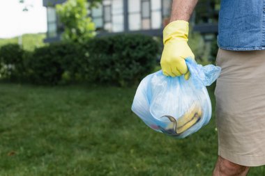 Cropped view of man in rubber glove holding bag with trash outdoors  clipart