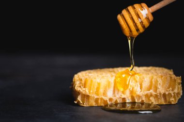 close up view of golden honey dripping on honeycomb from wooden dipper isolated on black clipart