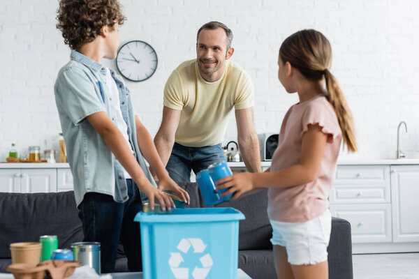 Smiling man looking at kids sorting trash near box with recycle sign at home 