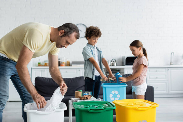 Man sorting garbage near blurred kids and trash cans with recycle sign at home 