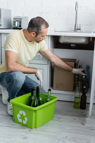 Man taking bottle near box with recycle sign in kitchen