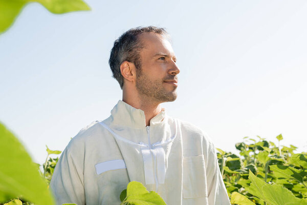 apiculturist in beekeeping suit looking away in field against clear sky, apiculture concept