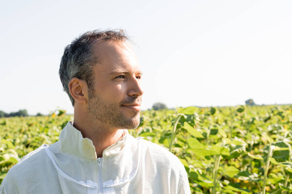 smiling bee master in beekeeping suit looking away in sunflowers field 