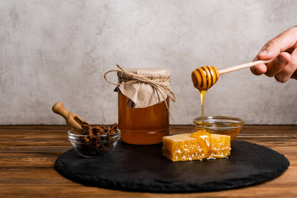 cropped view of man with wooden dipper near honeycomb, jar and bowls with honey and anise seeds on grey marble background