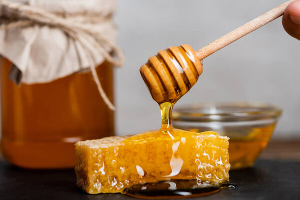 cropped view of male hand with wooden dipper near honeycomb and blurred honey jar isolated on grey