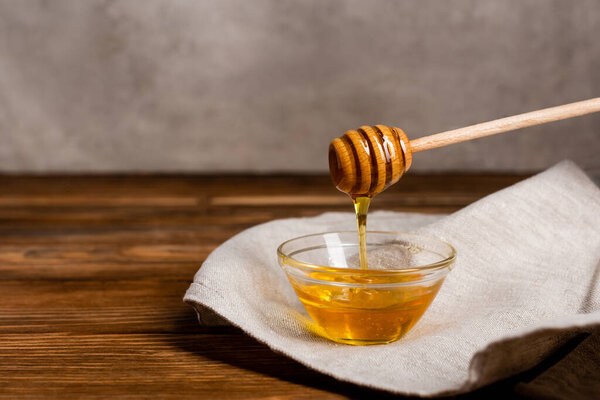 wooden dipper near bowl with liquid honey and linen napkin on wooden table and grey background