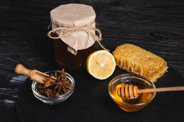 bowls with honey and anise seeds near fresh lemon, ginger root and jar covered with craft paper on black slate board