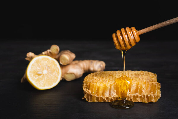 honey dripping from wooden dipper on honeycomb near blurred lemon and ginger root isolated on black