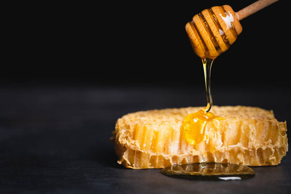 close up view of golden honey dripping on honeycomb from wooden dipper isolated on black