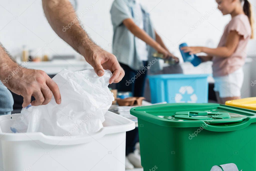 Cropped view of man holding plastic bag near blurred kids and trash cans at home 