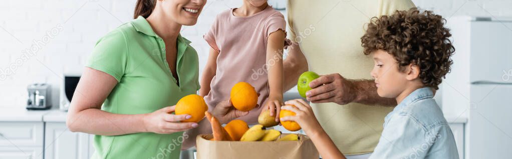 Smiling woman holding fruit near family and paper bag in kitchen, banner 
