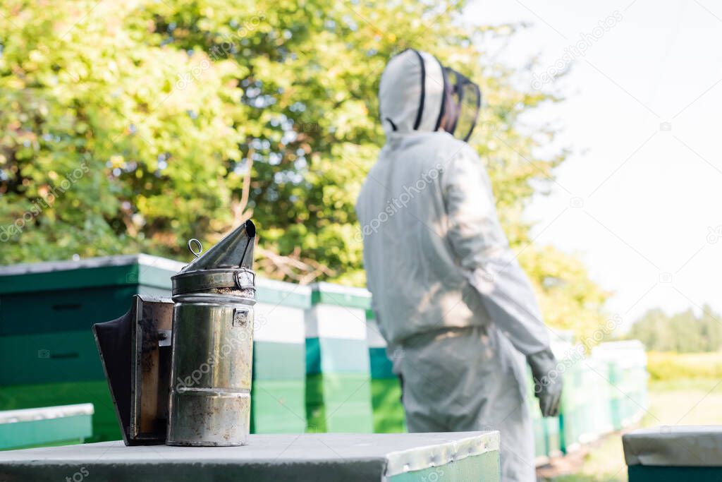 selective focus of bee smoker near beehives and blurred beekeeper in protective suit