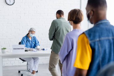 Tıp maskeli çok ırklı hastalar aşı merkezinde yazan olgun Asyalı doktorun yanında kuyrukta bekliyorlar.