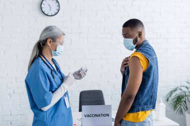 Tıbbi maskeli ve gözlüklü Asyalı doktor şırıngayı Afrika kökenli Amerikalı adamın yanında aşıyla dolduruyor.
