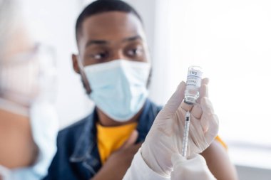 Lateks eldivenli hemşire Coronavirus aşısı ile dolduruyor. Tıbbi maskeli Afro-Amerikan bir adamın yanında.