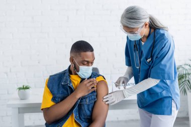 african american man in medical mask near asian nurse holding syringe with vaccine clipart