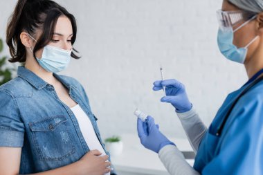 blurred asian doctor in medical mask and latex gloves showing jar with vaccine to pregnant woman clipart