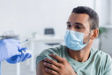 Doktor elinde şırınga ve aşı dolu kavanoz ile tıbbi maskeli genç bir adamın yanında.