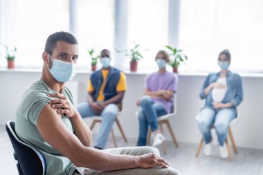 Koruyucu maskeli genç adam klinikte oturan çok ırklı insanların yakınındaki kameraya bakıyor, aşı konsepti.