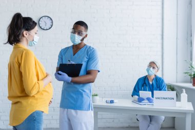 Hamile kadının yanında tıbbi maske takan Afrikalı Amerikalı doktor ve aşı merkezinde bulanık Asyalı meslektaşı.