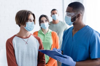 Tıbbi maskeli ve gözlüklü Afro-Amerikan doktor elinde pano, aşı merkezinde hastayla konuşuyor.