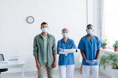 Çok ırklı doktorlar ve maskeli genç adam aşı merkezinde kameraya bakıyor.