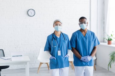 Maskeli ve gözlüklü ırklar arası doktorlar aşı merkezinde kameraya bakıyor.