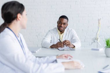 Gülümseyen Afrikalı Amerikalı doktor omurga modelinin yanında oturuyor ve bulanık meslektaşıyla konuşuyor.