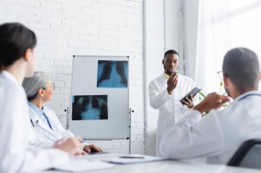 Afro-Amerikan doktor ciğerleri röntgenli bulanık meslektaşlarıyla konuşurken el kol hareketi yapıyor.