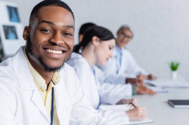 Genç Afrikalı Amerikalı doktor, arka plan bulanıklığı üzerine çalışan meslektaşlarının yanında kameraya gülümsüyor.