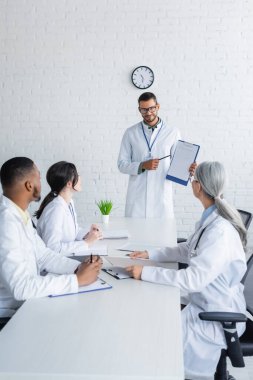 smiling doctor in eyeglasses pointing at clipboard near multiethnic colleagues sitting at desk clipart
