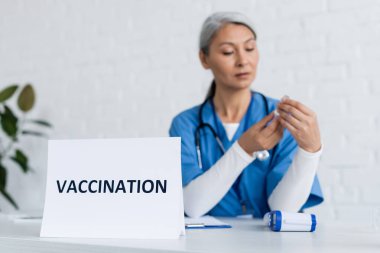 Olgun Asyalı doktor aşı dolu kavanozu kartın yanında aşı harfleriyle tutuyor.