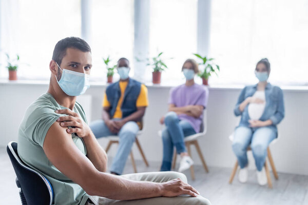 young man in protective mask looking at camera near blurred multiethnic people sitting in clinic, vaccination concept