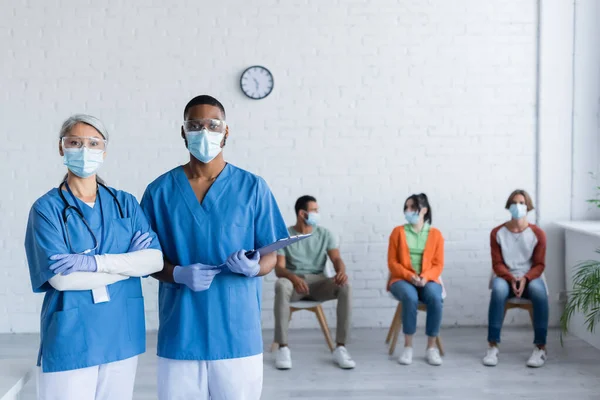 背景がぼやけている患者の近くのカメラを見ている医療用マスクやゴーグルの異人種間の医師 — ストック写真