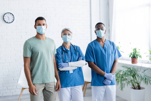 young man and interracial doctors in medical masks looking at camera in clinic, vaccination concept