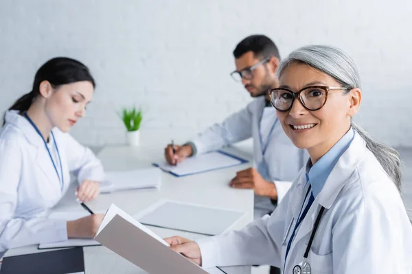Middle Aged Asian Doctor Eyeglasses Smiling Camera Young Colleagues Blurred — Stock Photo, Image