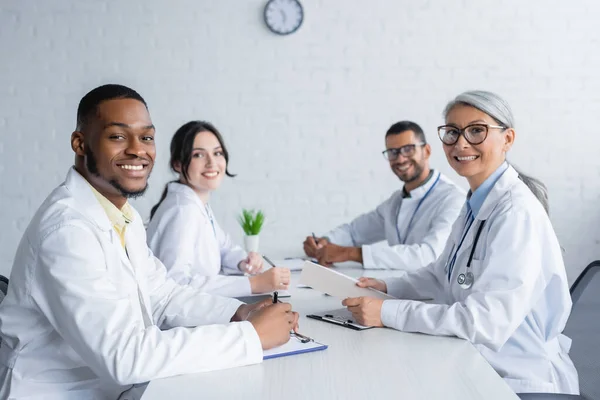 Médicos Multiétnicos Felices Mirando Cámara Mientras Están Sentados Escritorio Sala — Foto de Stock