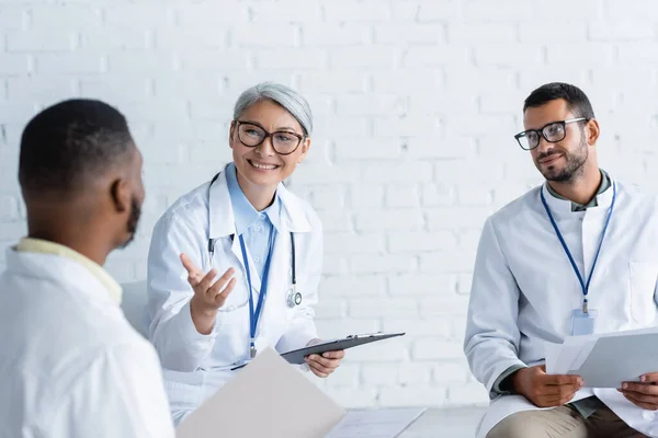 Sorrindo Asiático Médico Apontando Para Borrado Africano Colega Durante Discussão — Fotografia de Stock