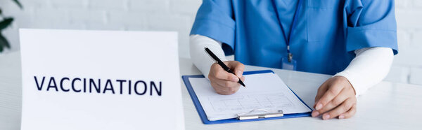partial view of immunologist writing on medical card near vaccination lettering on desk, banner