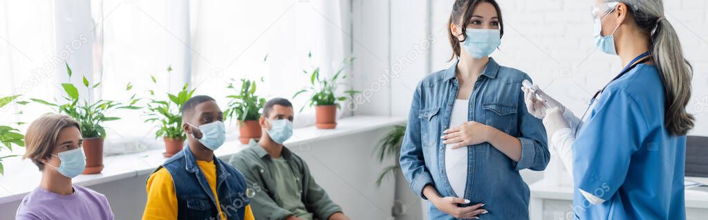 middle aged doctor in medical mask holding vaccine near pregnant woman and multiethnic patients, banner